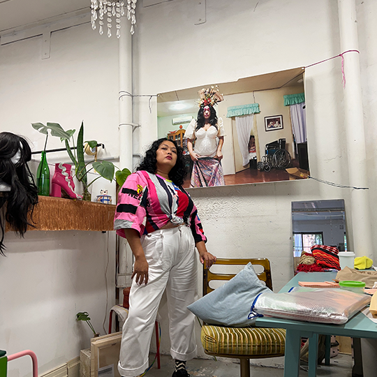 A photo of Rosell in her artists studio. Rosell wears white long pants and a abstract pink, white, grey, black, yellow, green and blue top, she stands infront of a photo of herself dressed as Ate Indai and leans on a chair in her studio. The studio has green plants, items of Ate's performance clothes, a pencil case, material, a mirror and a desk
